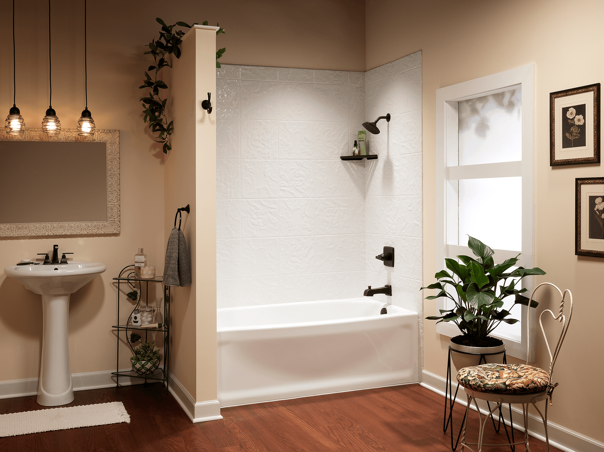 Warm, stylish bathroom with white tub and pedestal sink, pendant lights, framed art, leafy plants, and wood floors creates a calm retreat.