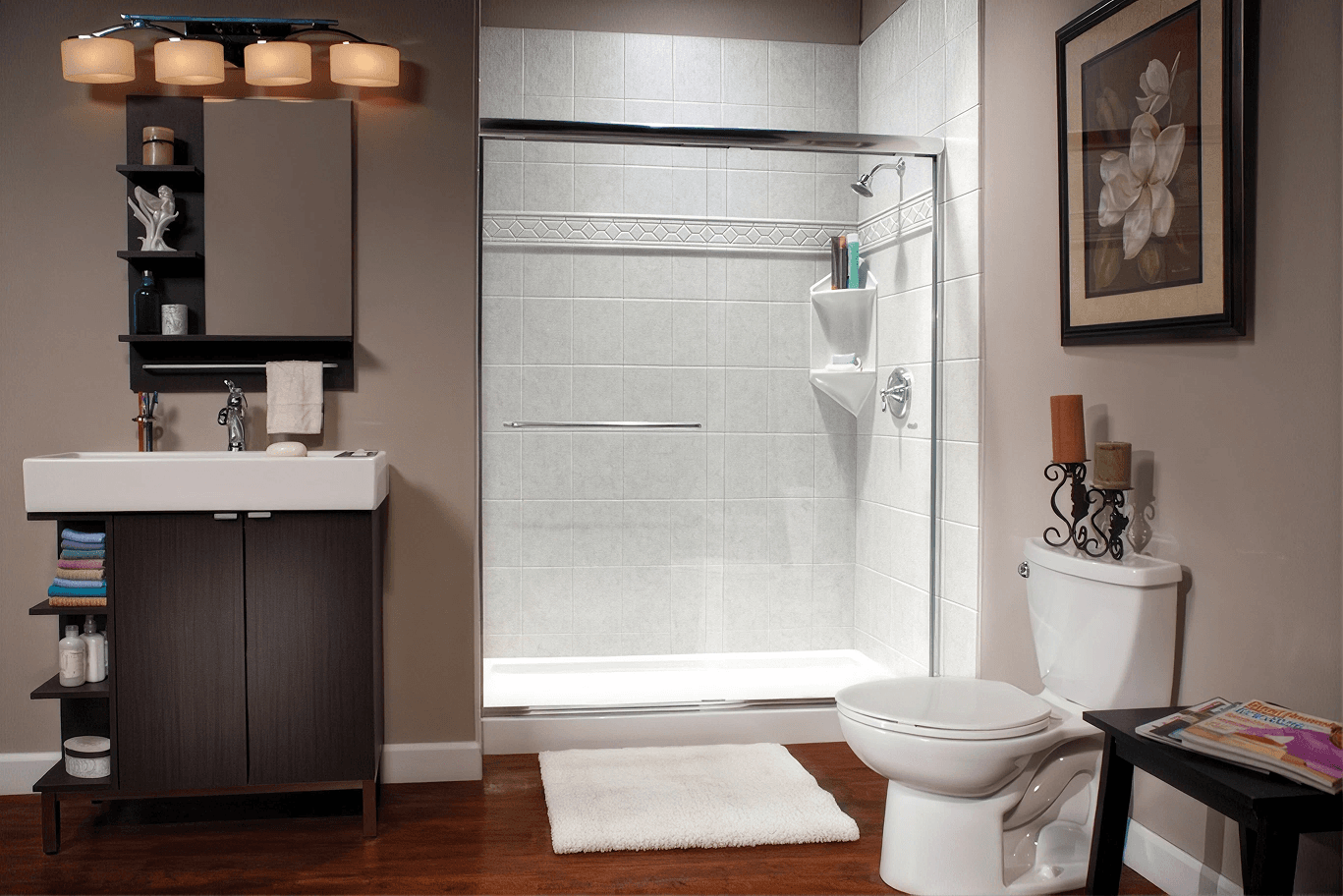 Modern bathroom with dark vanity, white sink, glass-enclosed shower with white tile surround, white toilet, and decorative accessories including framed botanical art and candles