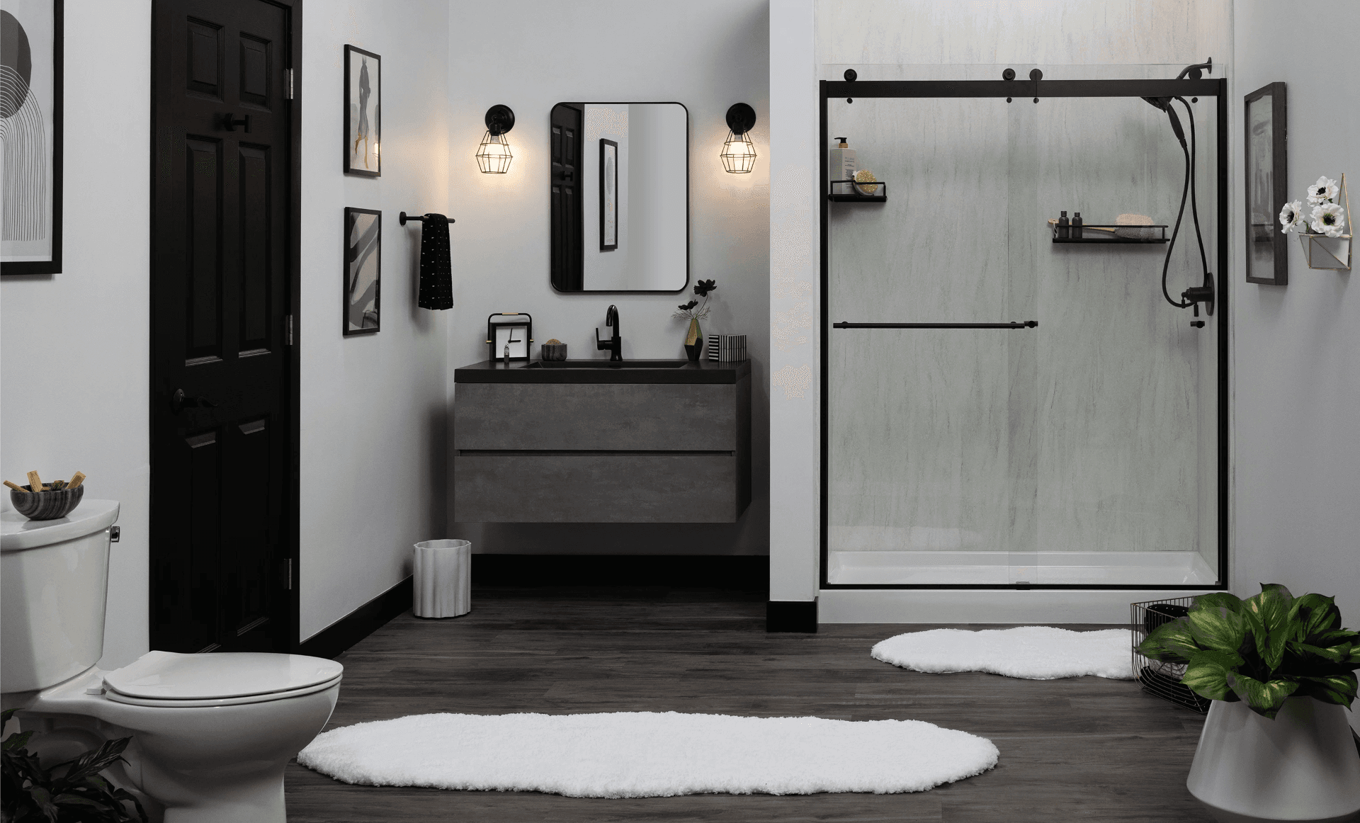 Modern bathroom with black and white color scheme, featuring a toilet, dark vanity with mirror and pendant lights, glass-enclosed shower with black frame, and white bath mats on dark wood flooring